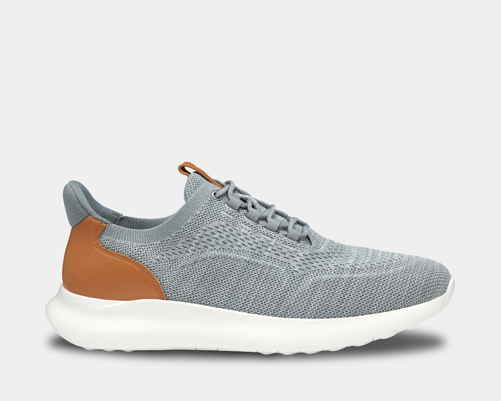 A pair of low-top athletic style knit sneakers with a plain toe design, featuring a white and gray heathered knit fabric, brown leather trim, and a tan heel counter.